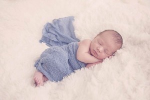 Newborn wrapped in a blue blanket laying on a white rug
